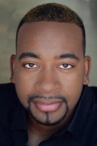 A close-up photo of a person with short, dark hair wearing a black shirt against a light-colored background.