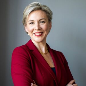 A white-presenting woman with short light blonde hair smiles as she wears a red jacket with her arms crossed.