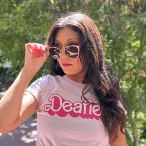 Leila outdoors wearing sunglasses and a pink “Deafie” T-shirt, holding the glasses while standing in sunlight.