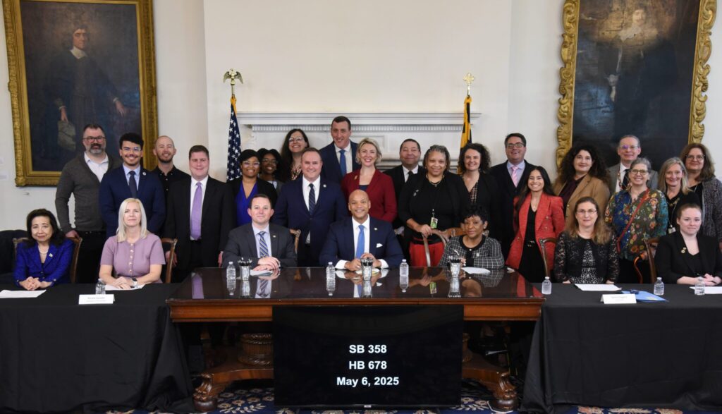 A large group of people pose together in a formal room with U.S. and Maryland flags behind them. Seven individuals are seated at a wooden table with a black sign reading “SB 358 HB 678 May 6, 2025.” 20 others stand behind.