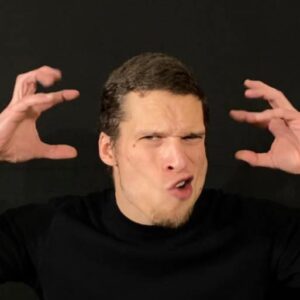 Man with a goatee wearing a black shirt makes an exaggerated angry expression with hands raised near his head.