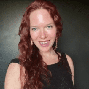Rach smiling with long curly red hair wearing a black top against a gray studio background.