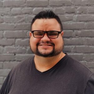 Justin smiling with short dark hair, beard, and glasses wearing a black T-shirt, standing in front of a gray brick wall.
