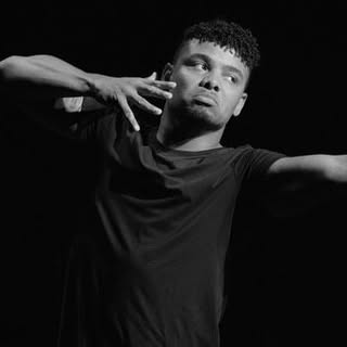 Black-and-white photo of Justin in a dark T-shirt, one arm extended out of frame and the other posed near his face against a black background.