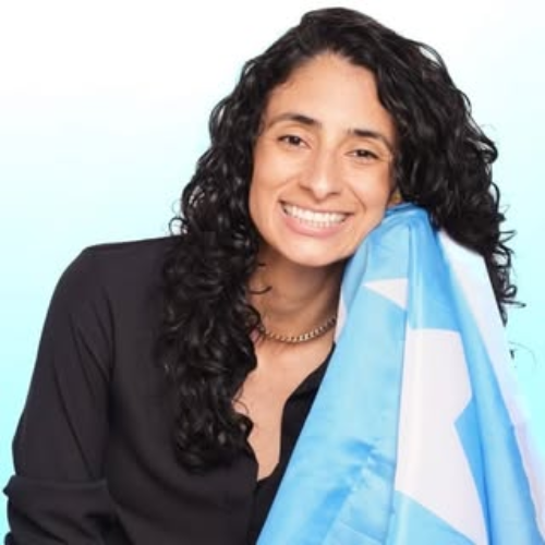 Smiling person with curly hair holds a light blue flag with a white star against a plain background.