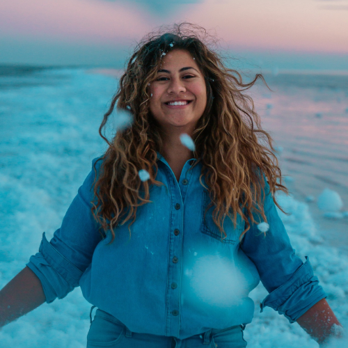 Person smiling in falling snow near a body of water at dusk, wearing a blue shirt.